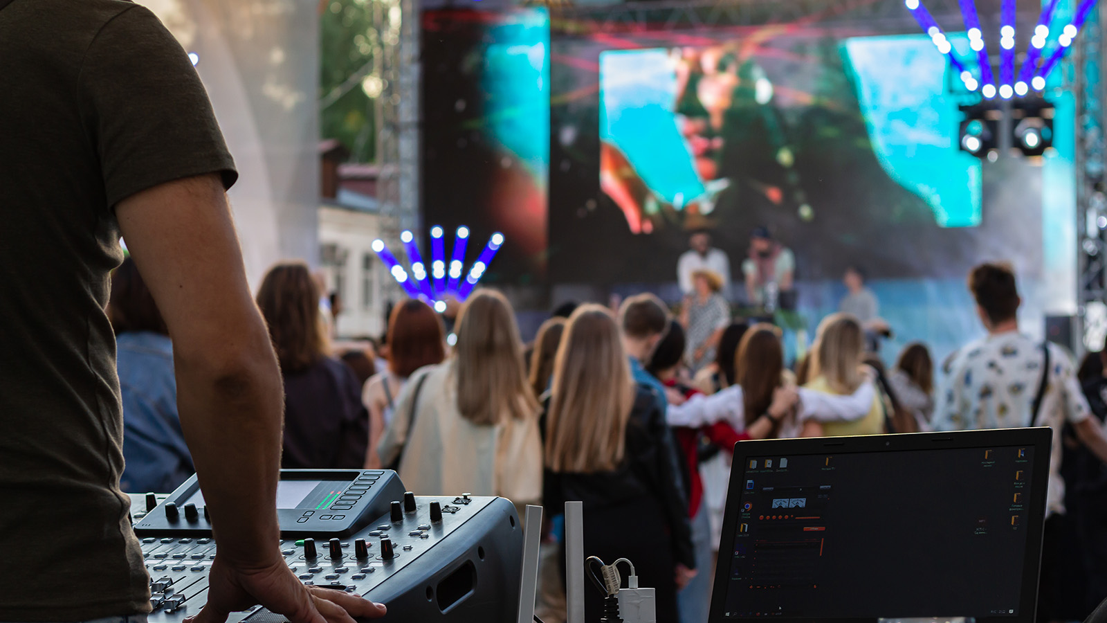 A sound technician stands next to a soundboard with his hand on the panel. Behind him is a large crowd of people and a stage with digital screens. The outdoor stage is setup with lights and decorations. The scene captures a vibrant atmosphere, a fun and engaging entertainment event.